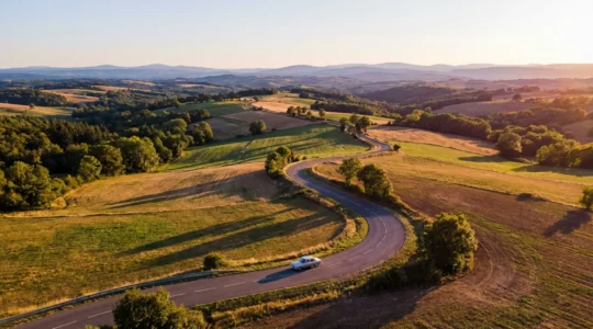 Vue aérienne d'une route de campagne sinueuse à travers les collines verdoyantes, avec une voiture sur une départementale pittoresque